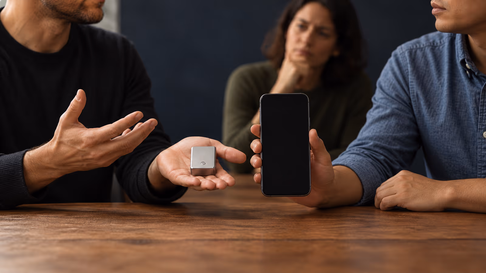In an imagined demonstration, a presenter holds out a small electronic device while a reviewer raises a smartphone beside it for comparison.